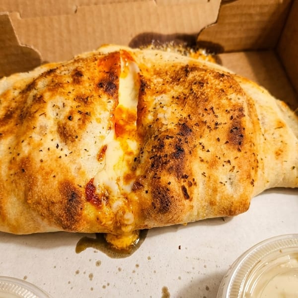 A golden-brown calzone with melted cheese and sauce oozing out, in a cardboard delivery box.