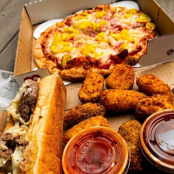 A pizza, cheesesteak, and fried appetizers with dipping sauces in takeout boxes on a wooden surface.